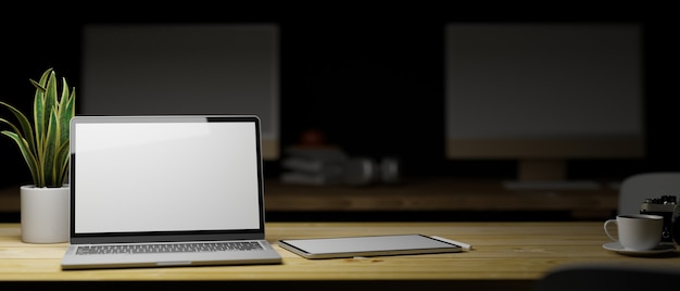 Pantalla en blanco del portátil con maqueta de tableta en la mesa de madera en el interior de la oficina con poca luz en segundo plano.