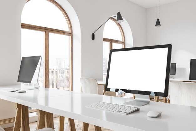 Pantalla de computadora blanca de pie en una mesa de oficina blanca en una habitación con ventanas arqueadas y paredes blancas.