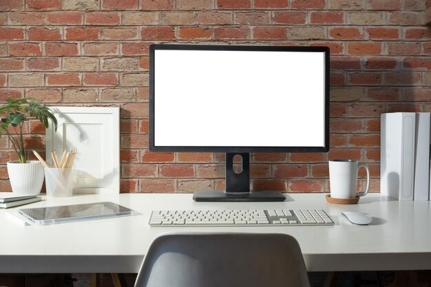 Pantalla de computadora de escritorio en blanco libros marco de imagen y planta de interior en mesa blanca contra pared de ladrillo