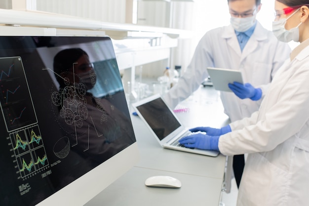 Pantalla de computadora con modelo de molécula en el fondo de dos trabajadores de laboratorio con batas blancas, máscaras y guantes que estudian información en línea