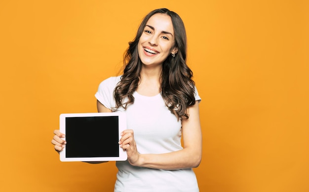Una pantalla de una tableta blanca que se coloca en las manos de una mujer agradable y feliz con una apariencia agradable y un atuendo informal fresco.