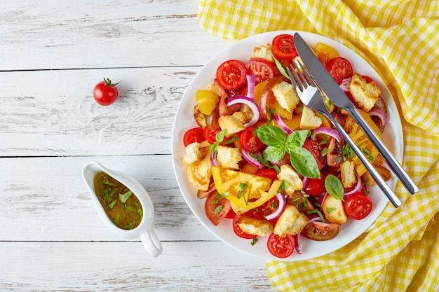 Panzanella deliciosa de salada italiana fresca com tomates, croutons e anéis de cebola em um prato branco sobre uma mesa de madeira com guardanapo, vista de cima, plana lay