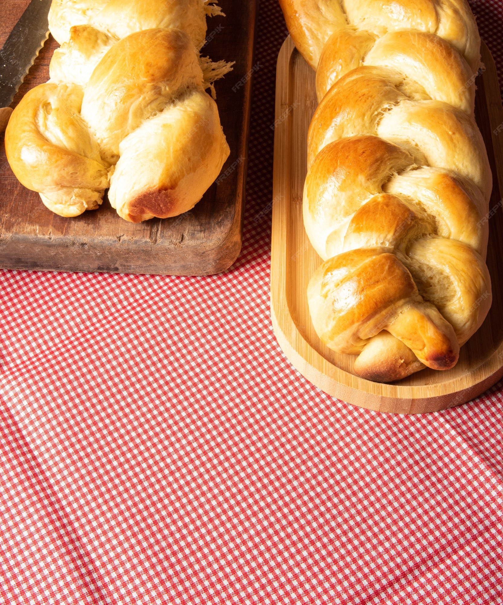 Pão caseiro, dois pães em forma de trança sobre madeira e uma toalha de  mesa quadriculada vermelha e branca, uma faca e um ramo de trigo.