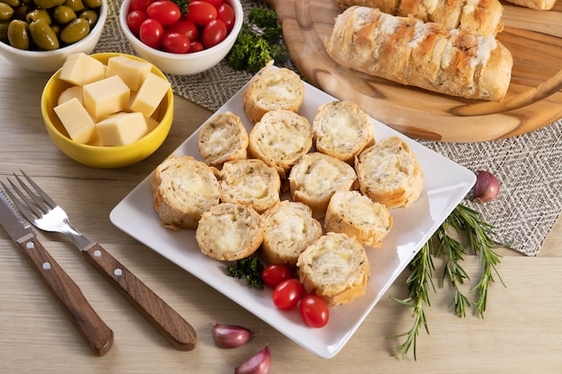 Pão de alho em prato quadrado branco em cima da mesa com queijo, alecrim, azeitonas e tomate cereja. Vista do topo.