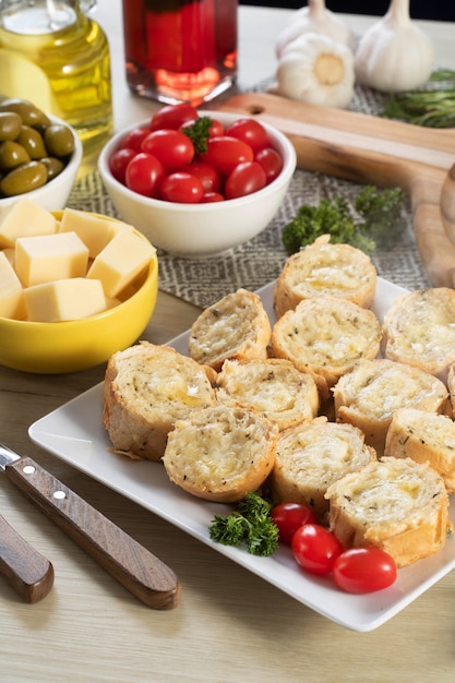 Pão de alho em prato quadrado branco em cima da mesa com queijo, alecrim, azeitonas e tomate cereja.