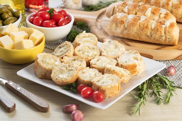 Pão de alho em prato quadrado branco em cima da mesa com queijo, alecrim, azeitonas e tomate cereja.