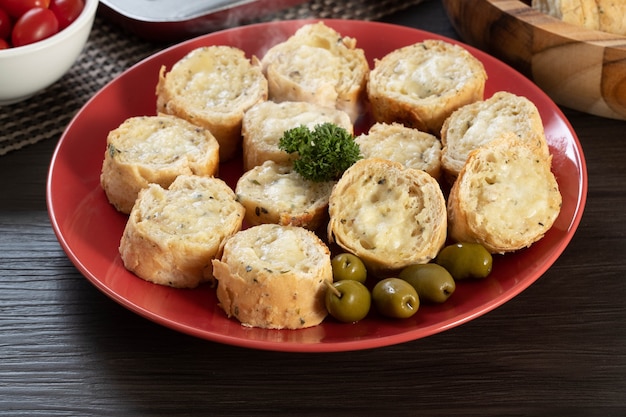 Pão de alho em um prato vermelho na mesa de churrasco com salsicha, queijo, alecrim, azeitonas e tomate cereja.