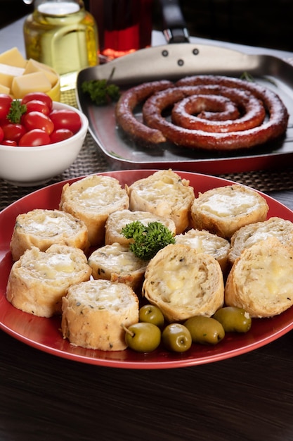 Pão de alho em um prato vermelho na mesa de churrasco com salsicha, queijo, alecrim, azeitonas e tomate cereja.