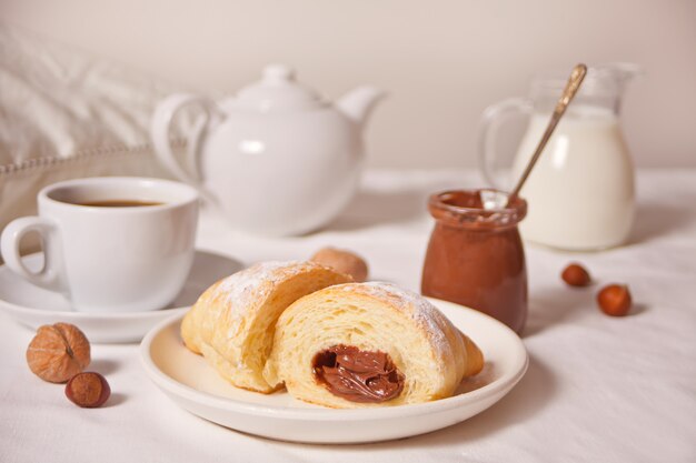 Pão de croissants frescos com chocolate no prato, xícara de café, jarra de leite