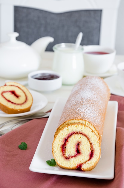 Foto pão de ló caseiro com geléia de framboesa em um prato branco sobre uma mesa de jantar de madeira branca com uma xícara de chá de baga perfumado.