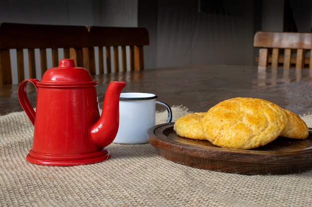 Pão de milho na placa de madeira, com bule vermelho e xícara branca na mesa