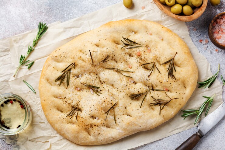 Pão focaccia caseiro tradicional italiano recém-assado no forno com temperos e alecrim em papel vegetal e fundo cinza claro. Vista do topo.