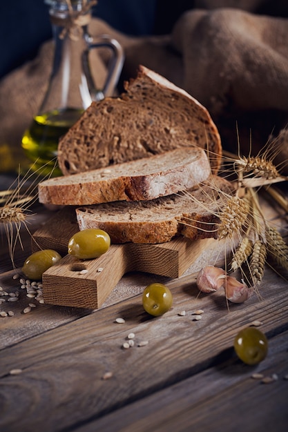 Pão fresco multigrãos crocante, azeitonas verdes e espigas de trigo em uma mesa de madeira rústica. Conceito de loja de alimentos de padaria e mercearia.