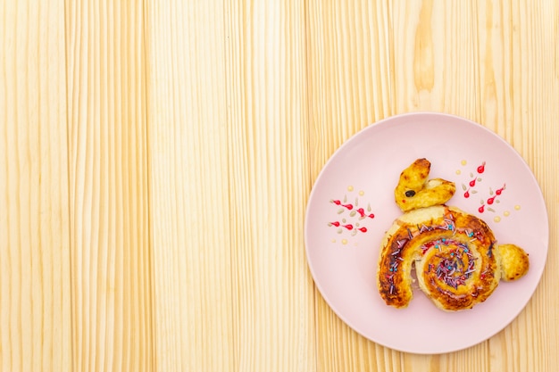 Pão fresco sob a forma de um coelhinho da Páscoa. O conceito de comida de férias infantil. Na placa cerâmica rosa na superfície de madeira, copie o espaço, vista superior.