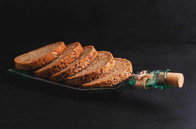 Pão wholegrain cortado com sementes de linho, sementes de girassol e sésamo em uma placa feita da garrafa no fundo preto.