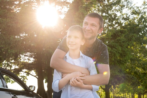 Foto papá abraza a un niño lindo de pie junto a un coche en el bosque contorno luz sol brillante