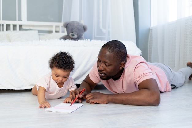 Papá afroamericano enseña al hijo de su hijo a dibujar con lápices en el suelo en casa