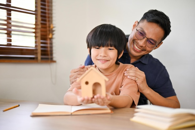 Papá asiático feliz abraza los hombros de su hijo mientras su hijo sostiene un modelo de casa