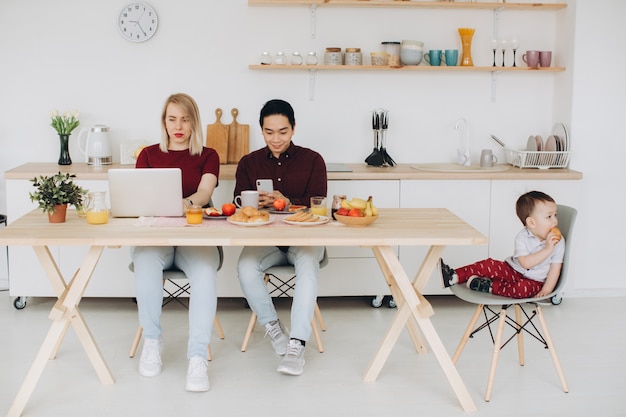 Papá asiático y mamá europea trabajan con sus gadgets, y su hijo desayuna solo. Problemas tecnológicos modernos.