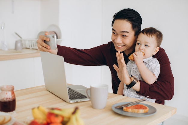 Papá asiático y su pequeño hijo desayunando en la computadora portátil y tomando selfie en el teléfono