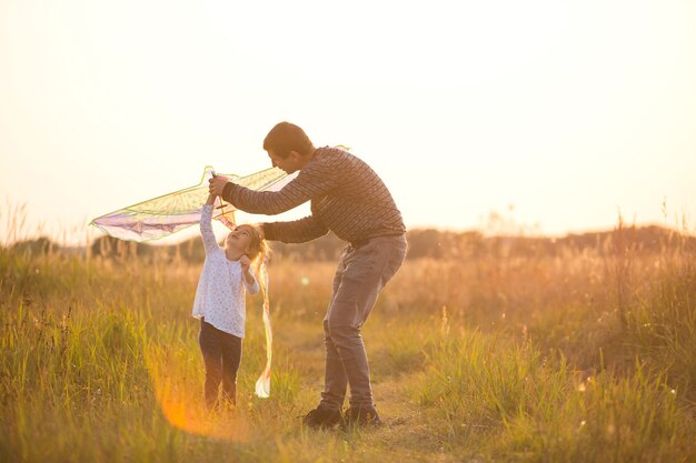 Papá ayuda a su hija a volar una cometa en un campo en el verano al atardecer. Animación familiar al aire libre, Día del Padre, Día del Niño. Zonas rurales, apoyo, asistencia mutua. Luz naranja del sol
