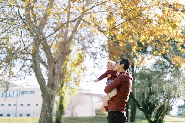 Foto papá besa a una niña en sus brazos en la mejilla mientras está de pie bajo un árbol con hojas amarillas