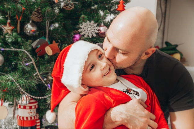 Papá besa a su hijo vestido con un disfraz de Papá Noel cerca del árbol de Navidad