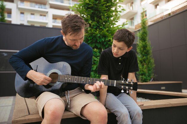 Papa bringt dem Sohn eines Teenagers bei, am Sommertag im Stadtpark Gitarre zu spielen, Konzept der gemeinsamen Interessen im Familienleben