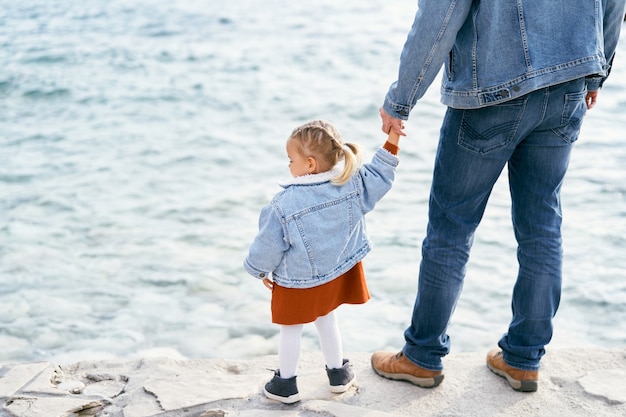 Papá en una chaqueta de mezclilla y jeans sostiene a una niña de la mano