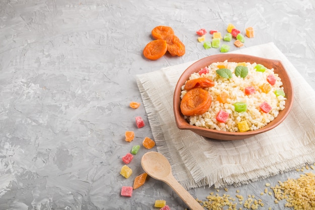 Papa de aveia de Bulgur com damascos secos e frutas cristalizadas na bacia da argila em um fundo cinzento concreto e em matéria têxtil de linho. Vista lateral, copie o espaço.