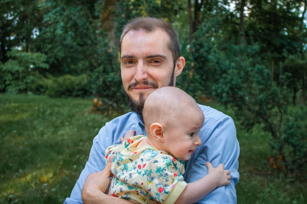 Papa, der seine Babysohnnahaufnahme auf dem Hintergrund einer Sommerlandschaft hält. Bärtiger Mann lächelnd und glücklich, Kamera mit verwirrtem Baby betrachtend