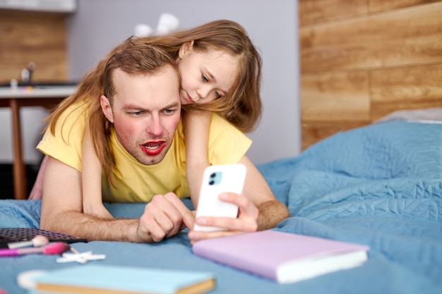 Papá e hija alegres tomando fotos en el teléfono inteligente mientras tienen maquillaje facial brillante