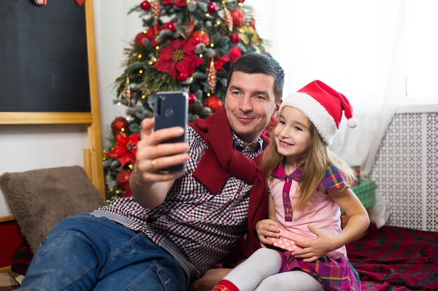 Papá e hija cerca del árbol de navidad con un teléfono inteligente se toman una selfie, se comunican a través de una conexión de video. saludos de navidad, una caja de regalo, una niña con un gorro de papá noel saludando con la mano.