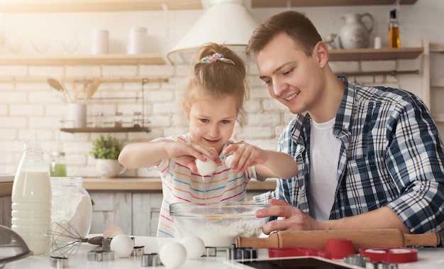 Papá e hija cocinando tarta sorpresa casera para mamá. La niña rompe el huevo de la masa en harina bajo la dirección del padre, copie el espacio