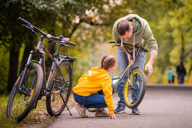 Papá e hija inspeccionan la rueda de la bicicleta adolescente para niños en el camino otoñal del parque