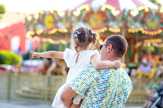 Papá e hija niña en un parque de diversiones en el verano de vacaciones ordenaron pasar tiempo, el niño señala con su dedo, vista trasera