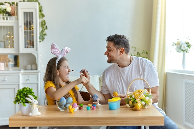 Papá e hija se tiñen la cara con pintura azul para pintar huevos. sobre la mesa hay una canasta con huevos de Pascua y pinturas.