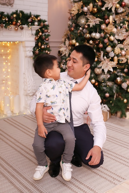 Papá e hijo asiáticos ríen, sonríen y se abrazan junto a la chimenea y el árbol de Navidad en casa