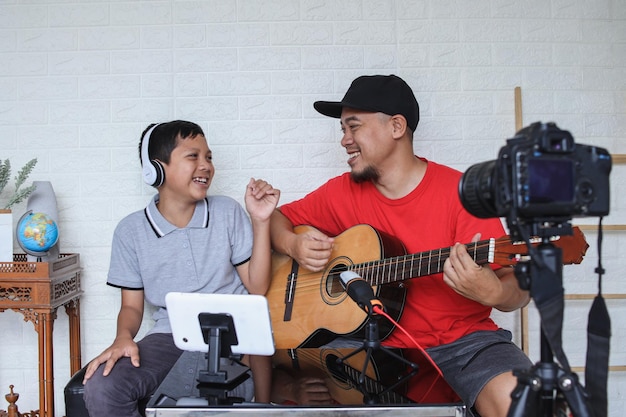 Papá e hijo cantan y tocan la guitarra juntos mientras hacen transmisión de video en línea. Familia asiática spe