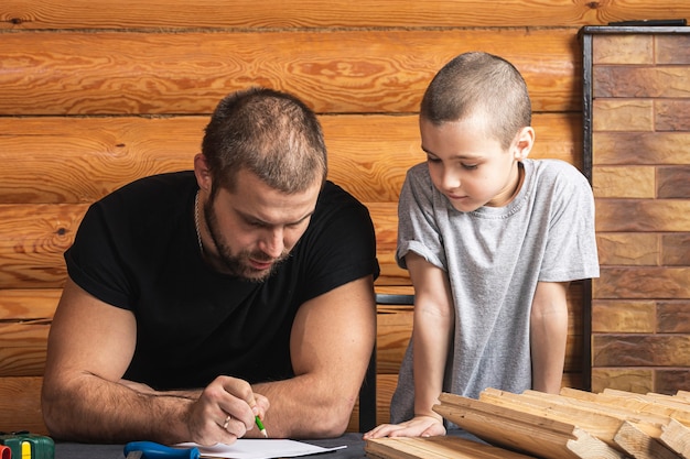 Foto papá e hijo dibujan en papel, planean cómo construir una casa para pájaros, herramientas y una viga en la mesa del taller.