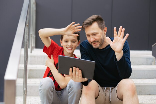 Papá e hijo hacen videollamadas con una tableta en la ciudad en una escalera de hormigón