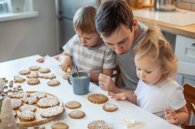 Papá e hijos decoran pan de jengibre navideño en casa un niño y una niña pintan con cornetas con sug ...