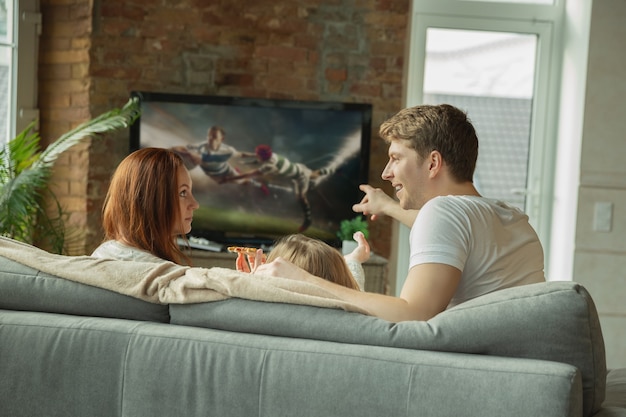 Foto papá eligiendo canal. familia pasando un buen rato juntos en casa. mamá, papá e hija divirtiéndose, comiendo pizza, viendo el campeonato de rugby en la televisión. unión, comodidad en el hogar, concepto de amor.