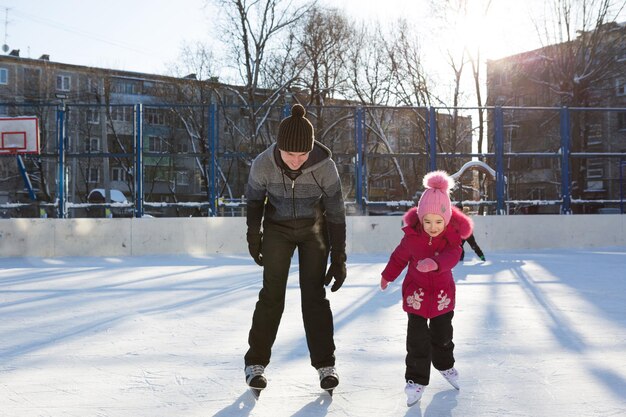 Papá le enseña a su pequeña hija a patinar sobre hielo en una pista de patinaje en el patio de los edificios de varios pisos de la ciudad. Día soleado de invierno helado, deportes de invierno activos y estilo de vida