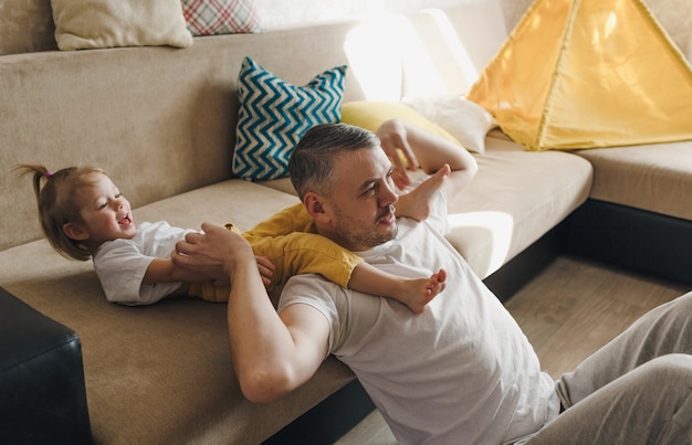 Un papá feliz con una camiseta blanca juega en el sofá con su hija, se ríe y disfruta pasar el tiempo con placer.