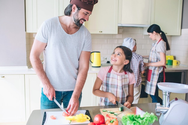 Papá feliz está con su hija y corta verduras. Se miran y sonríen. Mamá trabaja con el hijo detrás de ellos en la estufa.