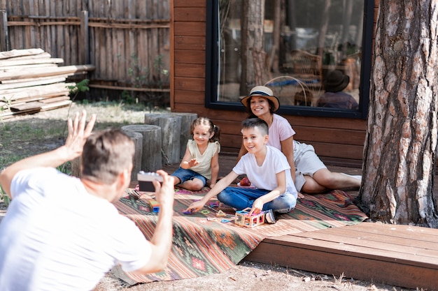 Papá fotografía a su esposa y sus dos hijos con una cámara de película, y la familia se divierte durante las vacaciones de verano. Sentado en una alfombra bordada en el porche cerca de una casa de madera.