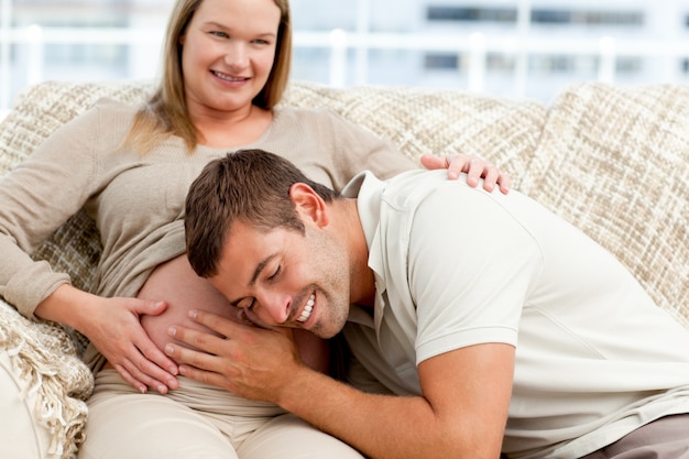 Papá futuro atento escuchando el vientre de su esposa sentada en el sofá