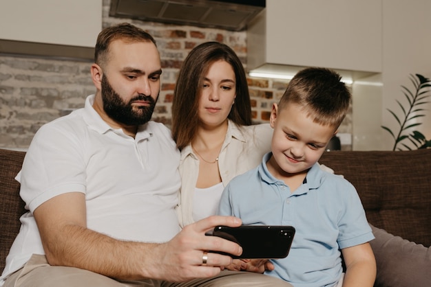 Un papá, un hijo y una mamá están viendo un video en el sofá. Un esposo sombrío con barba está demostrando el programa en el teléfono inteligente a un niño y una esposa por la noche en casa.