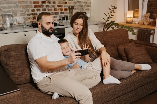 Un papá, un hijo y una mamá están viendo un video en un teléfono inteligente en el sofá. Un padre con barba está demostrando a su familia noticias en la pantalla del teléfono celular por la noche en casa acogedora.